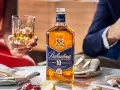 Elegancka kolacja z whisky i piwem w tle. Butelka Ballantine's 10 Years Old na stole, obok kieliszek z bursztynowym trunkiem.