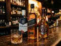 Trzy butelki blended whisky: Ballantine's, Johnnie Walker Black Label i Chivas Regal 12, stoją na barze obok szklanki z whisky i lodem.
