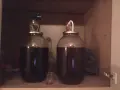 Dwa balony z ciemnym płynem, gotowe do dosłodzenia wina po fermentacji. Mały kieliszek obok sugeruje degustację.