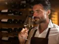 Mężczyzna wącha białe wino w kieliszku, zastanawiając się, w jakiej temperaturze robić wino. W tle stoją butelki w piwniczce.