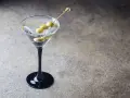 Elegancki martini z ginem, ozdobione trzema oliwkami na wykałaczce, w czarnej kieliszku na kamiennym tle.