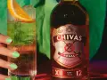 Chivas Regal 12 z czym pić? Orzeźwiający drink z plasterkiem pomarańczy i lodem obok butelki Chivas Regal 12.