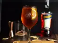 Orzeźwiający Aperol ze Sprite, udekorowany plasterkiem pomarańczy, na drewnianym blacie. Obok butelka Aperolu i inne akcesoria barmańskie.