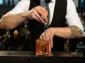 Barman miesza kostki lodu z bursztynowym płynem w szklance. Idealny **whisky z tonikiem** gotowy do podania.