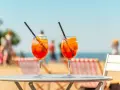 Dwa orzeźwiające drinki aperol z sokiem pomarańczowym, z lodem i plasterkiem pomarańczy, na tle plaży i błękitnego nieba. Idealne na wakacyjny relaks.