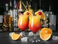 Dwa drinki z pomarańczą i wiśnią, ozdobione słomkami, na tle butelek i światełek.