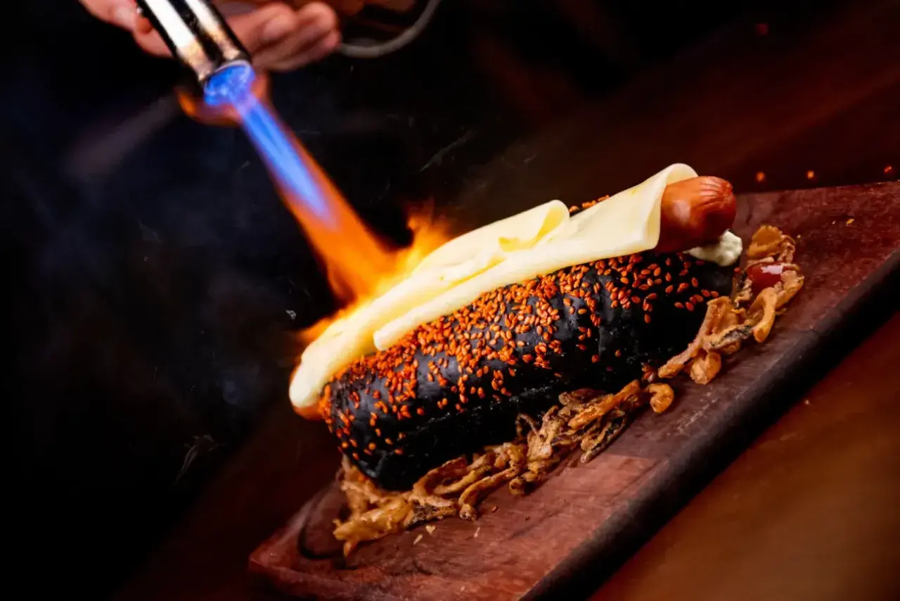 Czarny hot dog z serem i sezamem, podpalany palnikiem. To właśnie flambirowanie co to za technika!
