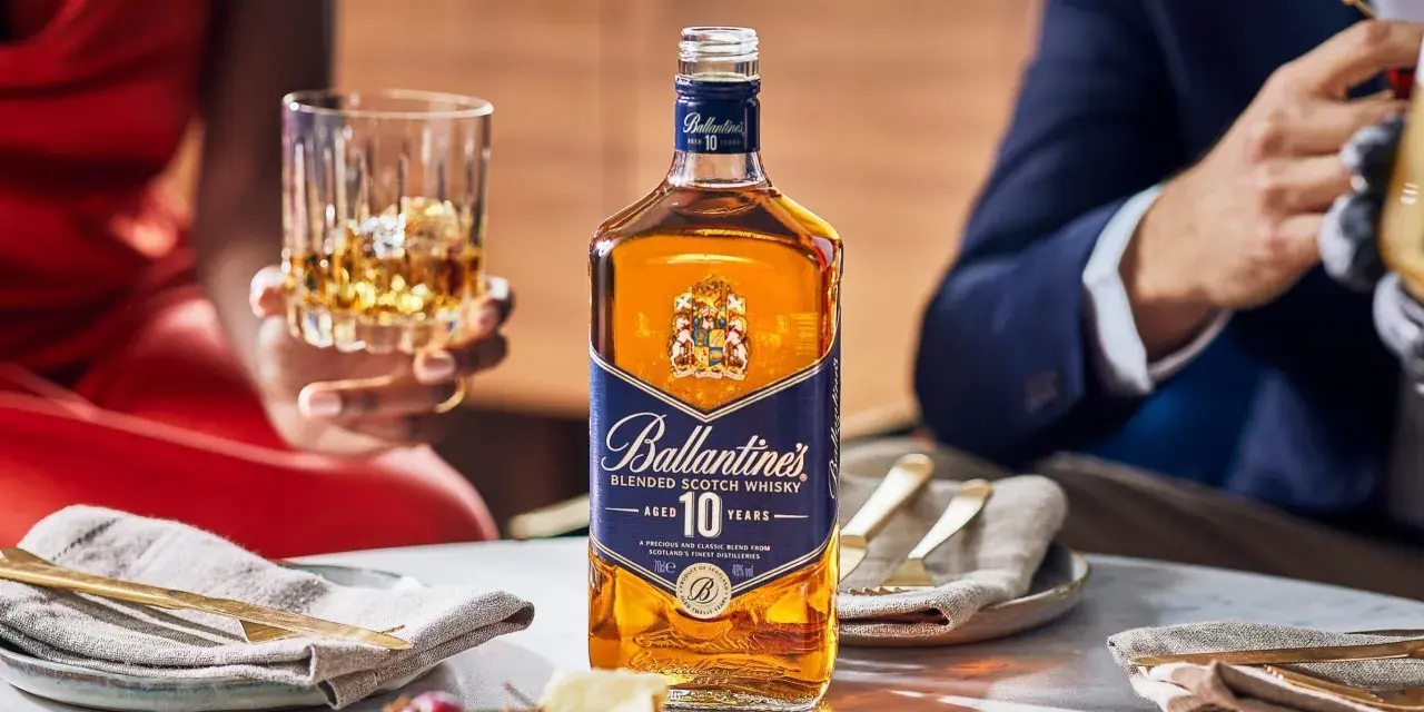 Elegancka kolacja z whisky i piwem w tle. Butelka Ballantine's 10 Years Old na stole, obok kieliszek z bursztynowym trunkiem.