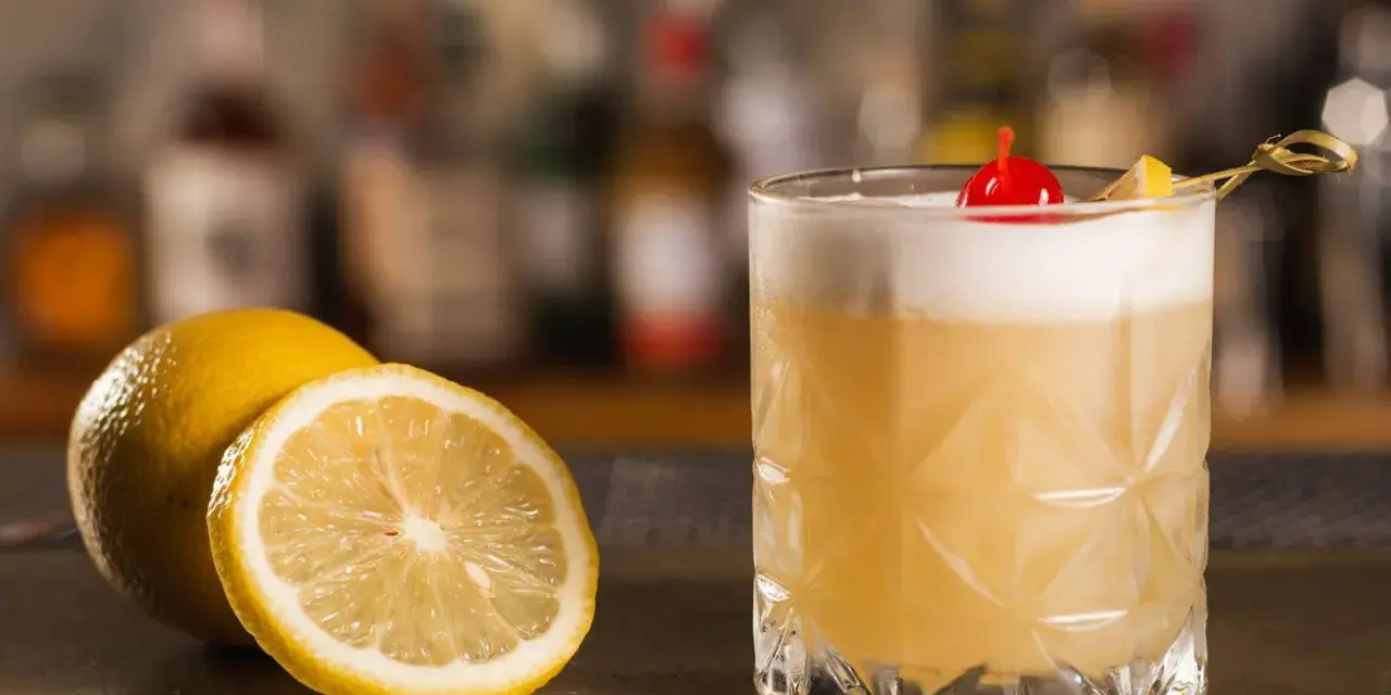 Orzeźwiający drink z amaretto, ozdobiony wisienką i plasterkiem cytryny, obok świeżych cytryn na barze.