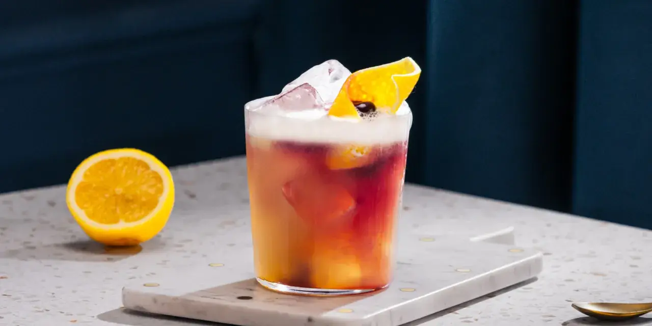 Orzeźwiający drink z whisky i cytryną, ozdobiony skórką cytrusową i lodem. Obok leży połówka cytryny.
