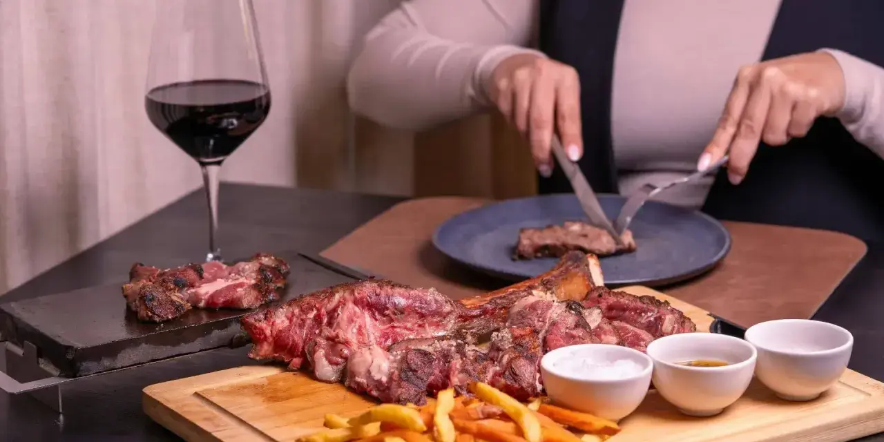 Idealne wino do dziczyzny: kieliszek czerwonego wina obok soczystego steka i frytek. Ktoś kroi mięso na talerzu.