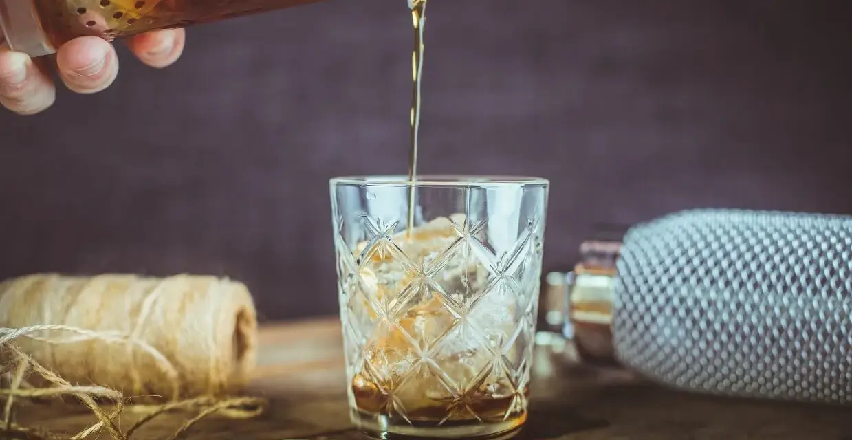 Nalewanie drinka z amaretto i wódką do szklanki z lodem. Na stole płatki migdałów i sznurek.