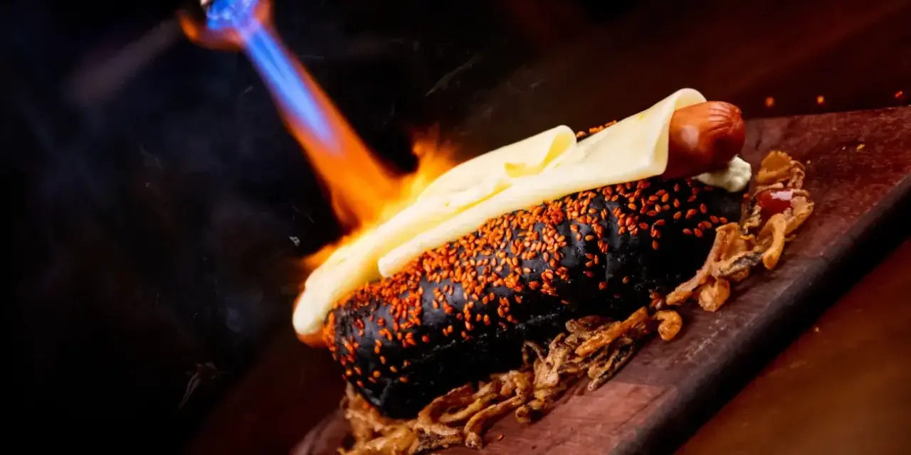Czarny hot dog z serem i sezamem, podpalany palnikiem. To właśnie flambirowanie co to za technika!