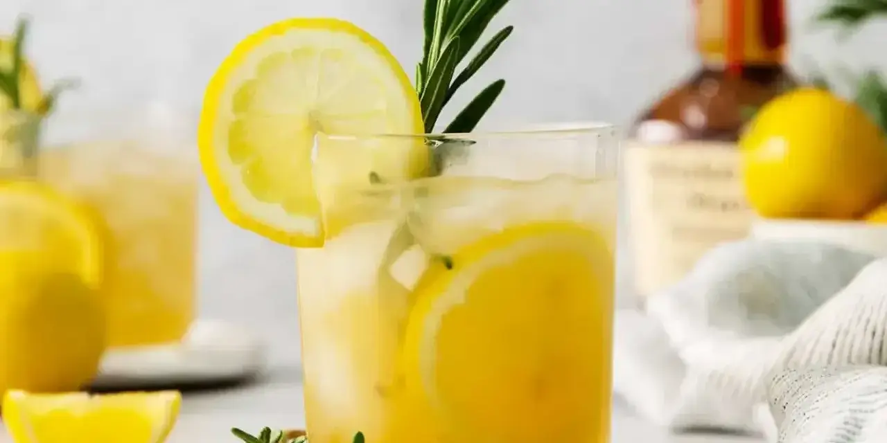 Szklanka z drinkiem z cytryną i rozmarynem, w tle butelka alkoholu i cytryny.