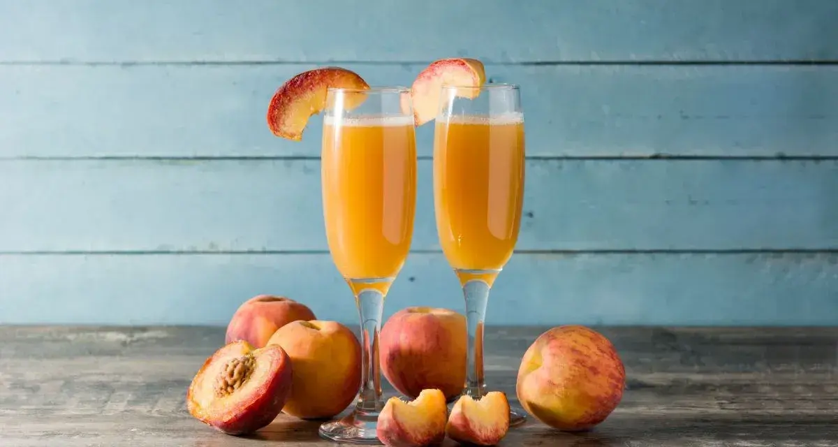 Dwa kieliszki z orzeźwiającym drinkiem na bazie likieru brzoskwiniowego, ozdobione plasterkami brzoskwini. Obok świeże owoce.