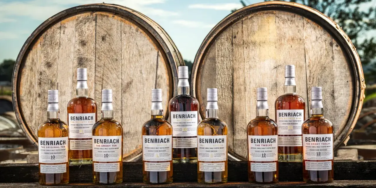 Butelki whisky BenRiach stoją przed beczkami, sugerując skąd pochodzi whisky.
