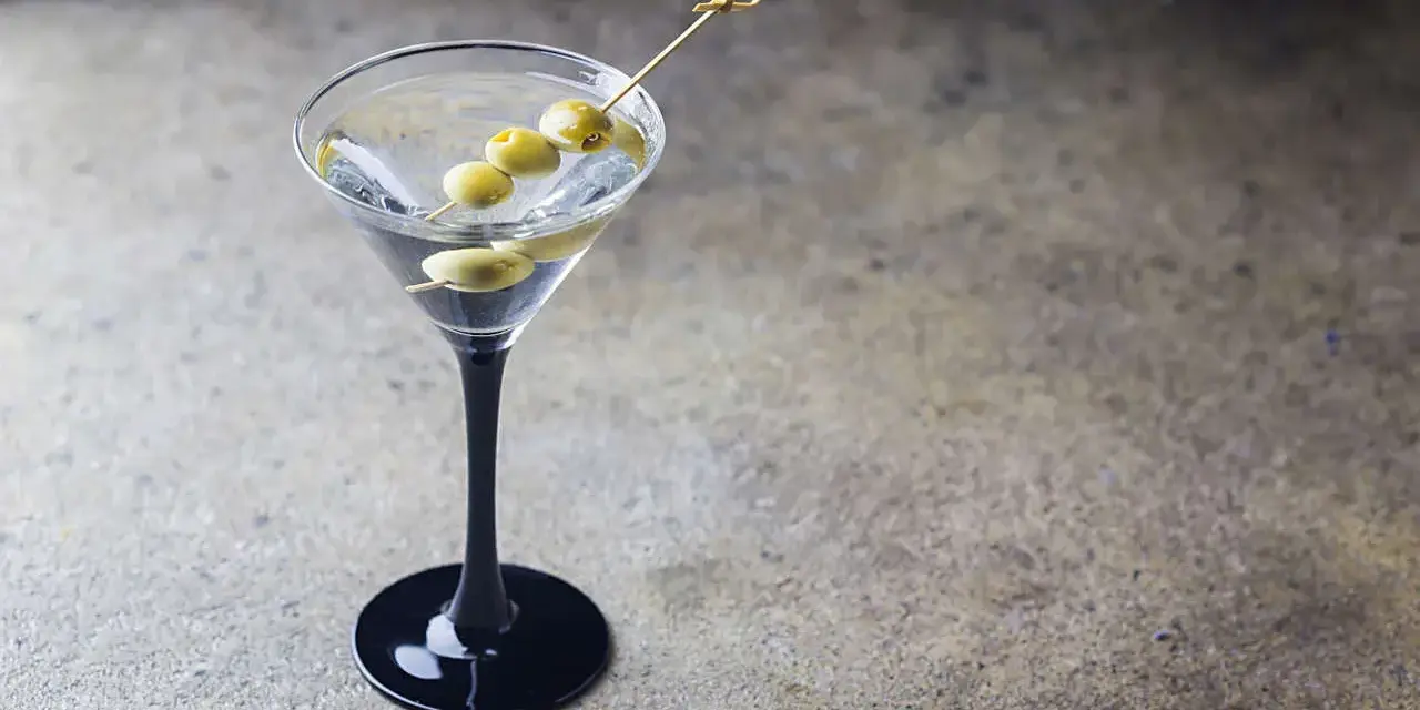 Elegancki martini z ginem, ozdobione trzema oliwkami na wykałaczce, w czarnej kieliszku na kamiennym tle.