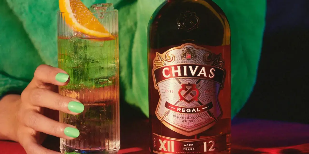 Chivas Regal 12 z czym pić? Orzeźwiający drink z plasterkiem pomarańczy i lodem obok butelki Chivas Regal 12.