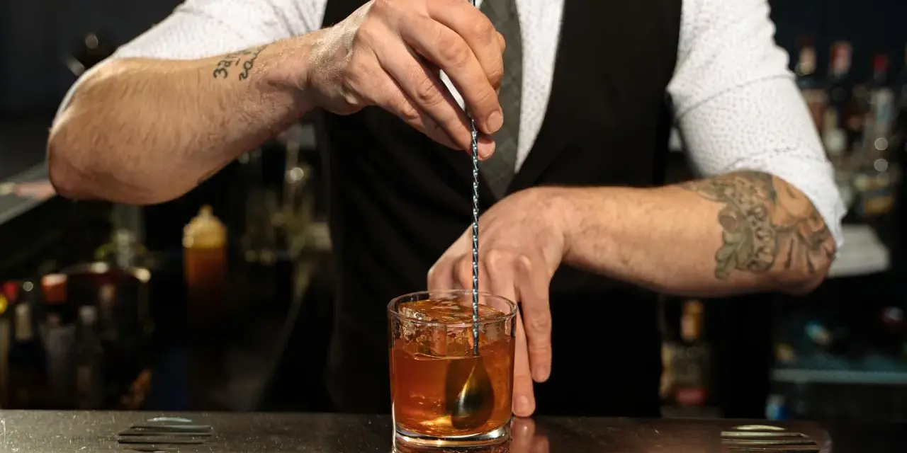 Barman miesza kostki lodu z bursztynowym płynem w szklance. Idealny **whisky z tonikiem** gotowy do podania.