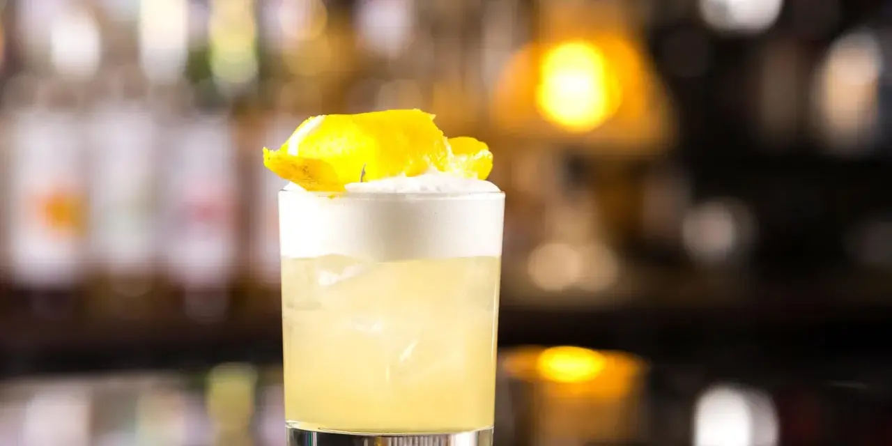 Orzeźwiający drink z whisky i miodem, ozdobiony skórką cytryny, na tle rozmytego baru.