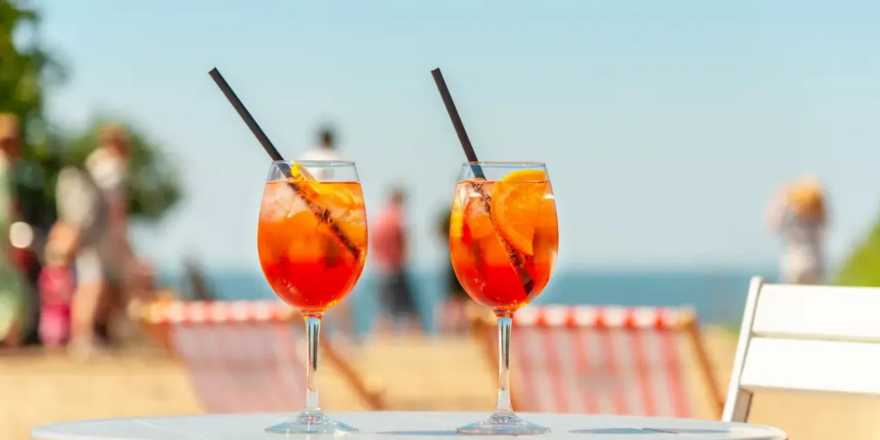 Dwa orzeźwiające drinki aperol z sokiem pomarańczowym, z lodem i plasterkiem pomarańczy, na tle plaży i błękitnego nieba. Idealne na wakacyjny relaks.