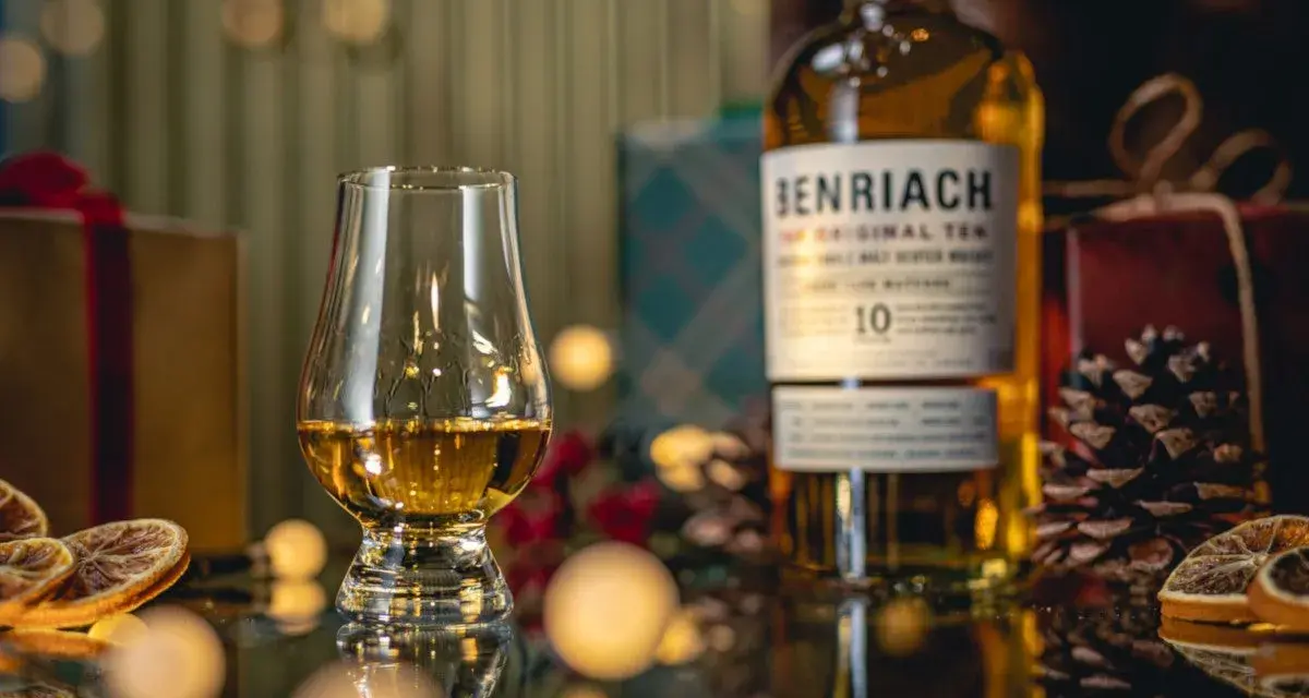 Szklanka z bursztynowym single malt whisky Benriach 10, obok butelki i świątecznych dekoracji.