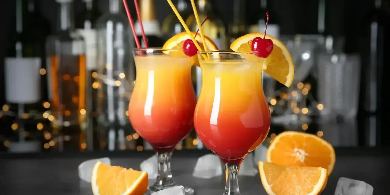 Dwa drinki z pomarańczą i wiśnią, ozdobione słomkami, na tle butelek i światełek.