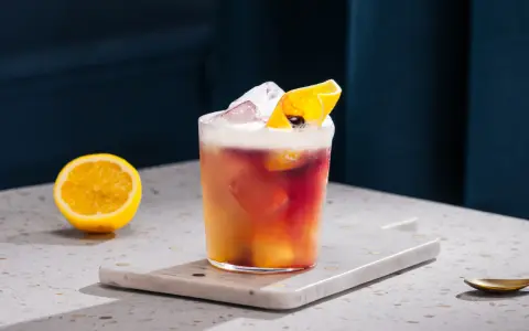 Orzeźwiający drink z whisky i cytryną, ozdobiony skórką cytrusową i lodem. Obok leży połówka cytryny.