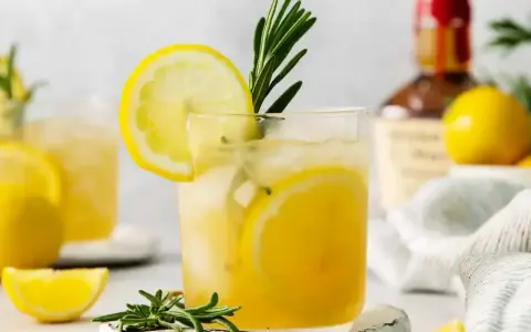 Szklanka z drinkiem z cytryną i rozmarynem, w tle butelka alkoholu i cytryny.