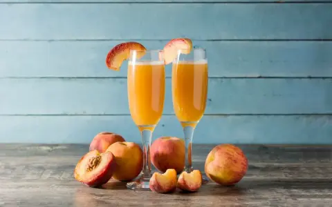 Dwa kieliszki z orzeźwiającym drinkiem na bazie likieru brzoskwiniowego, ozdobione plasterkami brzoskwini. Obok świeże owoce.