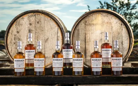 Butelki whisky BenRiach stoją przed beczkami, sugerując skąd pochodzi whisky.