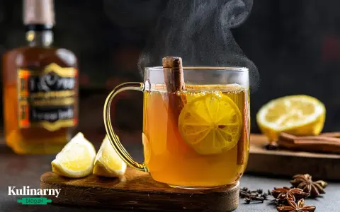Gorąca herbata z whisky, cytryną i cynamonem, parująca w szklance. Obok leżą cytryny i przyprawy.