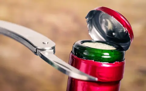 Metalowy widelec próbuje otworzyć butelkę wina, podważając czerwone wieczko.