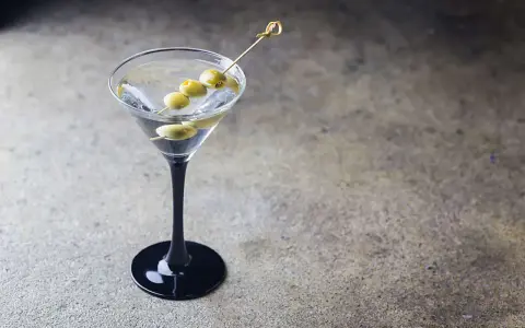 Elegancki martini z ginem, ozdobione trzema oliwkami na wykałaczce, w czarnej kieliszku na kamiennym tle.