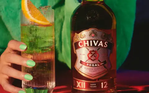 Chivas Regal 12 z czym pić? Orzeźwiający drink z plasterkiem pomarańczy i lodem obok butelki Chivas Regal 12.