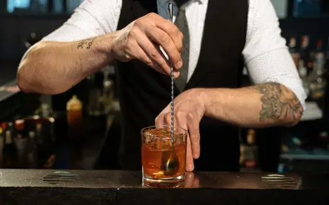 Barman miesza kostki lodu z bursztynowym płynem w szklance. Idealny **whisky z tonikiem** gotowy do podania.