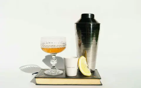 Elegancki kieliszek z bursztynowym trunkiem, shaker, plasterek cytryny i biały kubeczek z napisem "MIXING GLASS" – wszystko to sugeruje, z czym pić whisky, by stworzyć wyrafinowany koktajl.