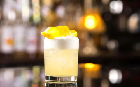 Orzeźwiający drink z whisky i miodem, ozdobiony skórką cytryny, na tle rozmytego baru.