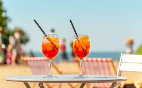 Dwa orzeźwiające drinki aperol z sokiem pomarańczowym, z lodem i plasterkiem pomarańczy, na tle plaży i błękitnego nieba. Idealne na wakacyjny relaks.