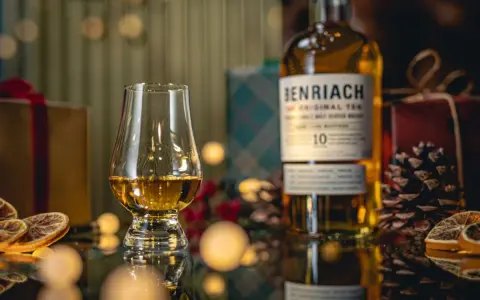 Szklanka z bursztynowym single malt whisky Benriach 10, obok butelki i świątecznych dekoracji.