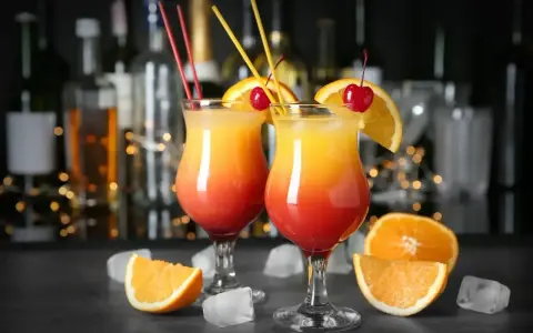 Dwa drinki z pomarańczą i wiśnią, ozdobione słomkami, na tle butelek i światełek.