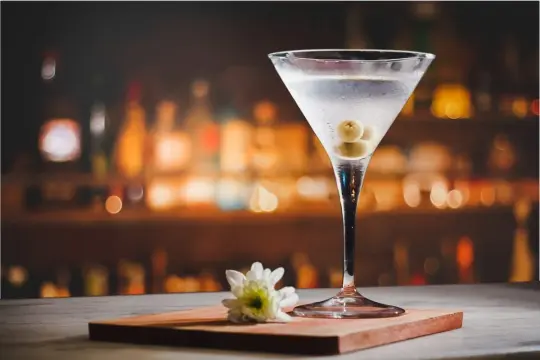 Kryształowy kieliszek martini z oliwkami, obok biały kwiatek. Idealne towarzystwo, by zastanowić się, z czym pić martini.