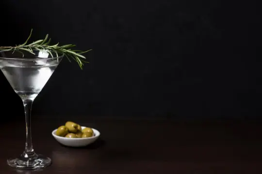 Elegancki martini z oliwką i gałązką rozmarynu, obok miseczka z zielonymi oliwkami.