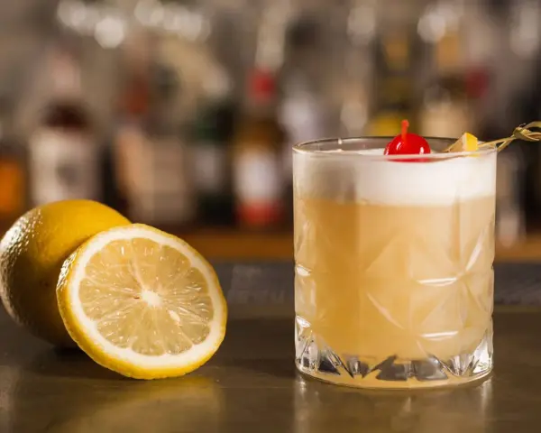 Orzeźwiający drink z amaretto, ozdobiony wisienką i plasterkiem cytryny, obok świeżych cytryn na barze.