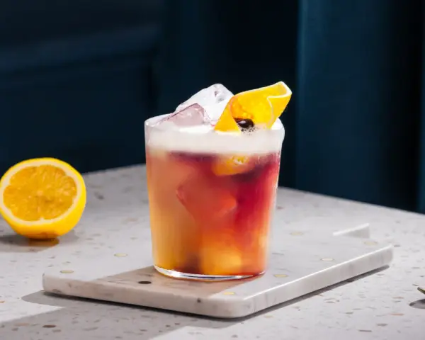 Orzeźwiający drink z whisky i cytryną, ozdobiony skórką cytrusową i lodem. Obok leży połówka cytryny.