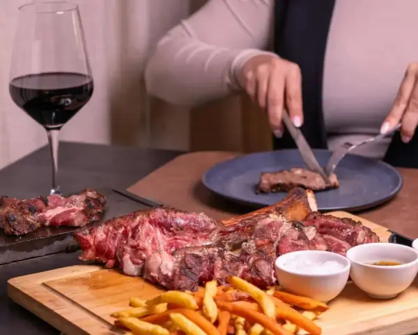 Idealne wino do dziczyzny: kieliszek czerwonego wina obok soczystego steka i frytek. Ktoś kroi mięso na talerzu.