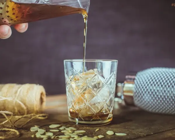 Nalewanie drinka z amaretto i wódką do szklanki z lodem. Na stole płatki migdałów i sznurek.