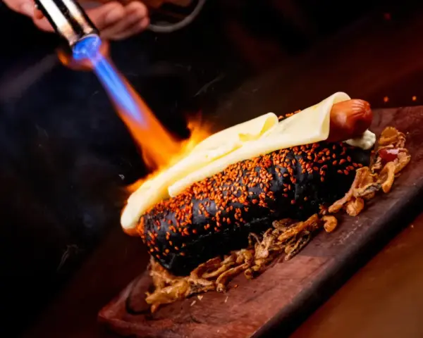 Czarny hot dog z serem i sezamem, podpalany palnikiem. To właśnie flambirowanie co to za technika!