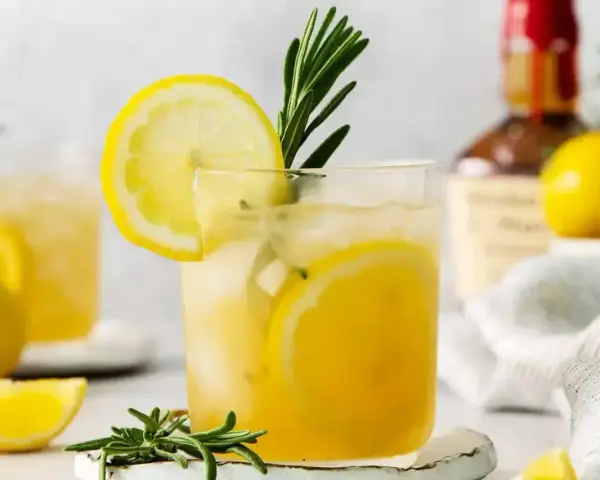 Szklanka z drinkiem z cytryną i rozmarynem, w tle butelka alkoholu i cytryny.