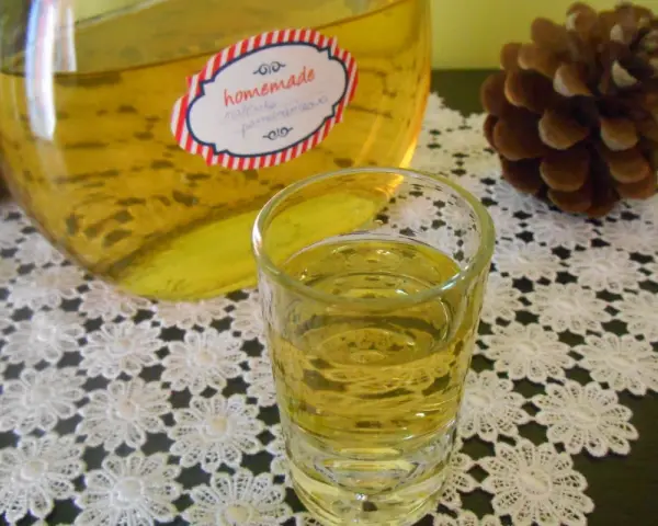 Szklaneczka i butelka z domową nalewką z mandarynek na koronkowym obrusie, obok szyszki.
